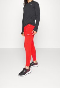 Zwarte top met lange mouwen met een klein Nike-logo, gecombineerd met strakke rode broek met zijrits zakken en een opvallend wit Nike-logo. Zwarte sneakers.
