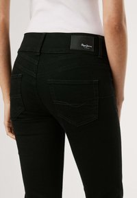 Prikaz blizu osobe koja nosi crne traperice s vidljivim stražnjim džepovima i oznakom Pepe Jeans London na pojasu, u kombinaciji s bijelom majicom.