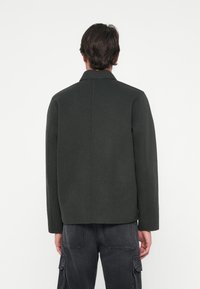 Mørkegrønn ulljakke med en strukturert silhuett, med høy krage og minimalistisk design. Bak har den en enkelt søm som går vertikalt.