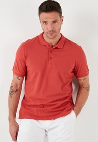 Polo tričko s krátkým rukávem v barvě spálené oranžové, vyrobené z hladkého materiálu. Má rovný lem a zapínání na tři knoflíky.