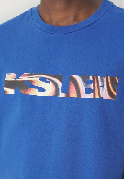 Camiseta de algodón azul con un estampado gráfico que presenta el texto "KSUBI" en patrones coloridos y en espiral. Manga corta y diseño de cuello redondo.