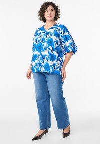 Blumenbluse in Weiß und Blau, mit V-Ausschnitt und Puffärmeln, kombiniert mit blauen Jeans im geraden Schnitt und schwarzen High Heels.