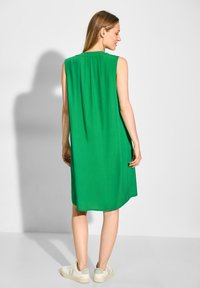 Robe verte sans manches, longueur genoux, avec une coupe décontractée et un pli dans le dos, associée à des baskets de couleur claire arborant un logo discret.