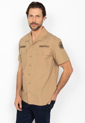 Homme barbu portant une chemise beige à manches courtes avec des poches avant et des écussons, debout avec une main dans la poche.
