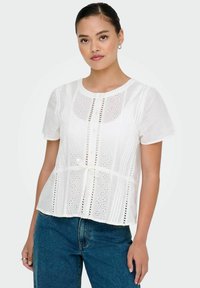 Weiße kurzärmelige Bluse mit floralem Stickerei, Lochmuster-Details, rundem Ausschnitt und einer Schleife in der Taille; kombiniert mit blauen Denim-Jeans.