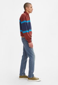 Camisa de polo de manga larga en burdeos con rayas horizontales azules y marinas, combinada con jeans azul claro y zapatillas verde olivo.