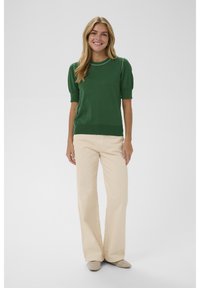 Mladá žena se usmívá, má na sobě zelený svetr s krátkým rukávem, béžové kalhoty s širokými nohavicemi a béžové boty se cvočky, stojí na jednolitém světlém pozadí.