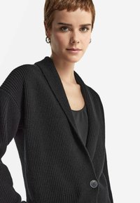 Svart ribbad kofta med sjalkrage, långa ärmar och en enda knappstängning. Tillverkad av ett texturerat stickat tyg.