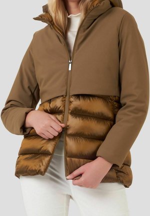Donna che indossa una giacca marrone con cappuccio e parte inferiore imbottita, che sta chiudendo la cerniera, vestita con pantaloni bianchi e una maglietta chiara.