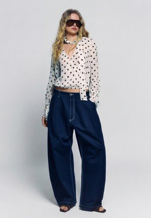 Frau trägt übergroße dunkle Sonnenbrille, weiße Bluse mit schwarzen Punkten, weit geschnittene dunkelblaue Jeans und schwarze Schuhe mit offenen Zehen.