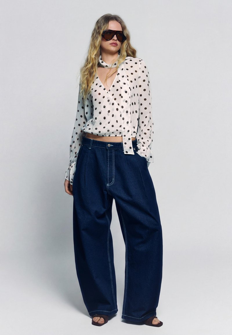 Žena nosi veľké tmavé slnečné okuliare, bielu blúzku s čiernymi bodkami, široké tmavomodré rifle s voľnými nohavicami a čierne otvorené topánky.