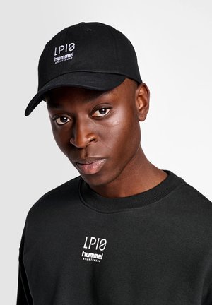 Ung mand iført en sort kasket og sweatshirt, begge med hvide "LP10 hummel SPORTSWEAR" logoer, mod en ensartet lys baggrund.