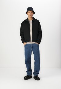 Veste zippée noire, t-shirt gris, jeans bleus et chaussures noires épaisses ; un modèle portant un chapeau seau noir se tient contre un fond blanc.