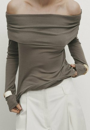 Woman wearing an olive off-shoulder long-sleeve top with thumb holes paired with high-waisted white pleated pants.