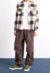 Rutig flanellskjorta i kräm, grå och orange; vit t-shirt under; bruna vida byxor; gröna mockaskor med vita sulor.