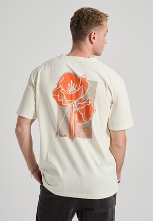 Man draagt een crème T-shirt met een grote oranje en beige bloemafbeelding en tekst op de rug, staand tegen een lichtgrijze achtergrond.