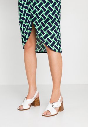 Onderbenen met witte sandalen met houten blokhakken en een marineblauwe rok met een groene en witte geometrische print en een split aan de voorkant.