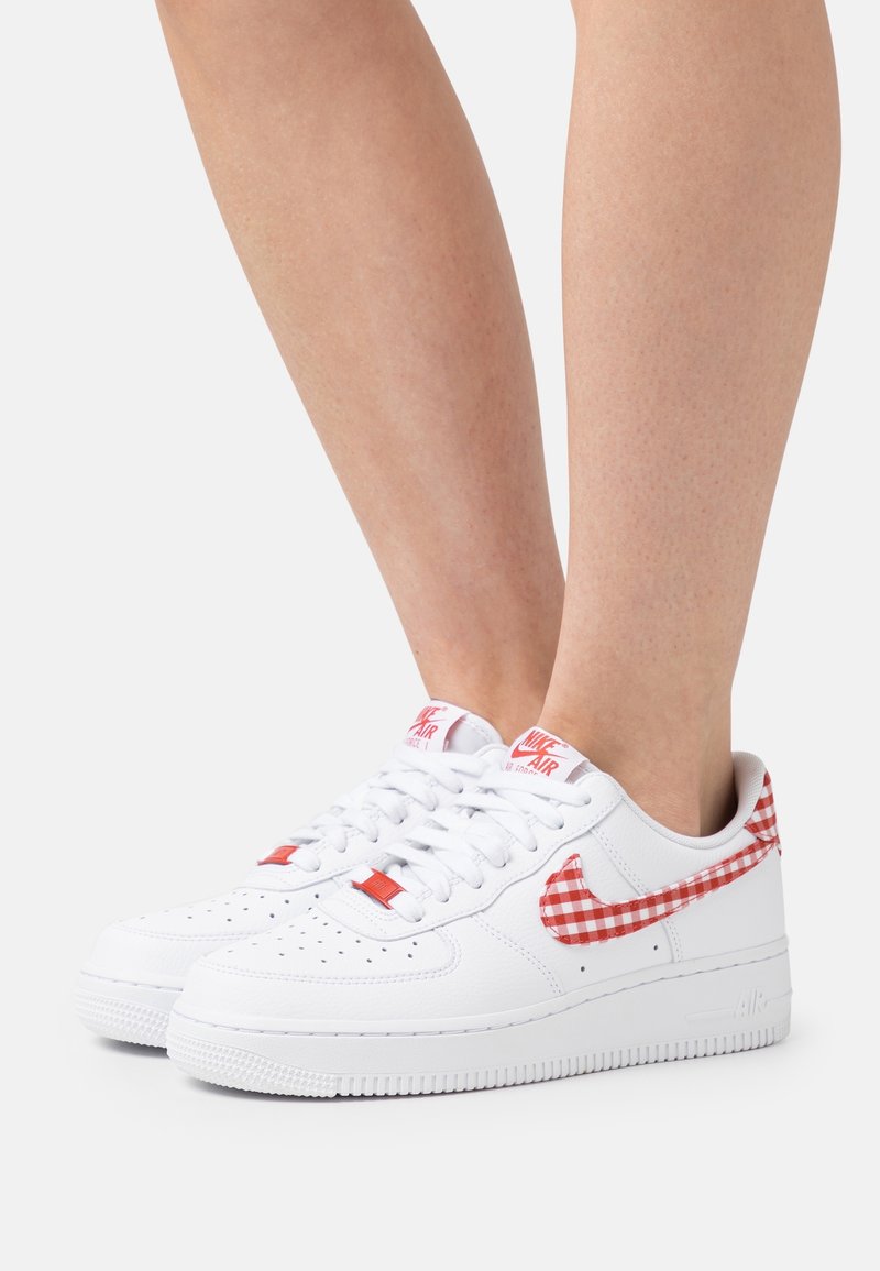 Baskets blanches avec une finition en cuir lisse, motif vichy rouge sur le Swoosh, et accents rouges sur la languette et les lacets.