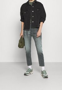 Svart denimjacka med fransar, grön stickad topp, urtvättade blå jeans och gröna/vita sneakers med texturerade detaljer. Håller en grön väska.