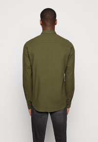 Uomo visto di spalle che indossa una camicia a maniche lunghe verde oliva e jeans grigio scuro su uno sfondo chiaro e neutro.