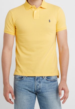 Homme portant un polo jaune à manches courtes avec un petit logo brodé et un jean bleu clair sur un fond uni.