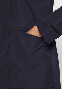 Main d'une personne insérée dans la poche zippée d'une veste bleu marine ; la manche comprend une sangle velcro noire réglable.