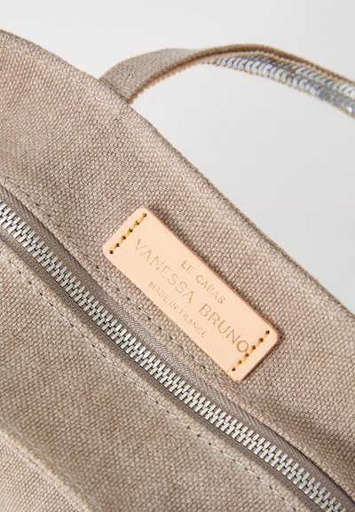 Sac en toile beige avec une fermeture éclair en métal visible, étiquette en cuir portant le nom "Vanessa Bruno", et des détails cousus. Texture lisse et design simple.