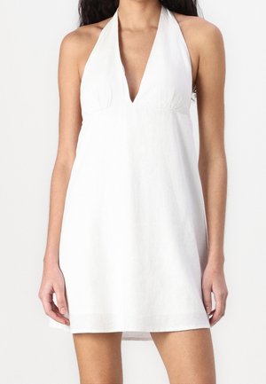 Femme portant une robe blanche sans manches avec un décolleté en V profond, debout devant un fond blanc uni.