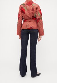 Quiltet jakke i støvet rosa med rød blomsterbroderi, bælte i taljen og lange ærmer, parret med mørke flared jeans og sorte flade sko.
