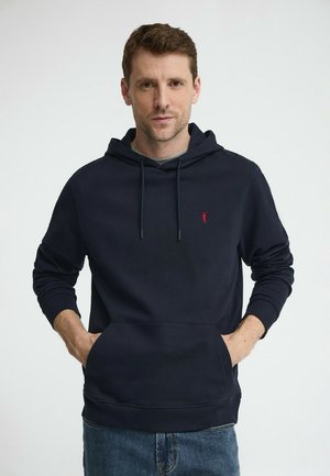 Mężczyzna w granatowej bluzie z kapturem z przednią kieszenią i małym czerwonym logo, stojący na tle białej ściany z rękami w kieszeniach.