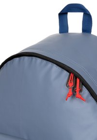 Un sac à dos bleu avec une forme arrondie, un matériau texturé, une fermeture éclair noire et des accents de cordon rouges. Doté d'une poignée bleue robuste sur le dessus.