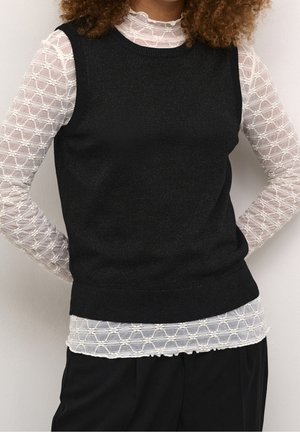Personne portant un pull sans manches noir sur un haut blanc en dentelle à manches longues avec un motif géométrique et un pantalon noir.