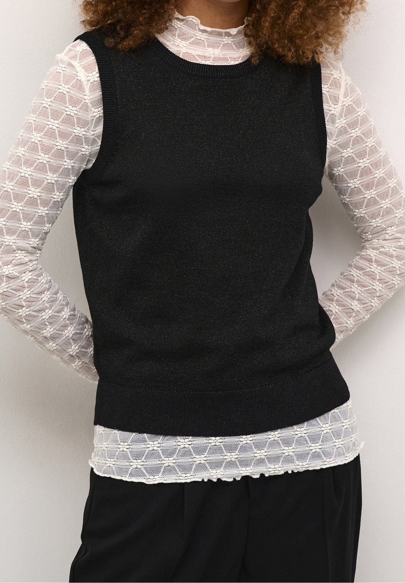 Personne portant un pull sans manches noir sur un haut blanc en dentelle à manches longues avec un motif géométrique et un pantalon noir.