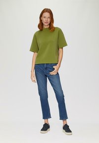 Olivenfarbene Kurzarmbluse, lockere Passform. Kombiniert mit blauen gerade geschnittenen Jeans und schwarzen Sneakers mit cremefarbenen Sohlen. Schlichter, minimalistischer Stil.