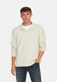 Polo de manga larga color beige claro con una textura suave, que presenta un cuello con solapa y un dobladillo recto. Combinado con jeans azul oscuro.