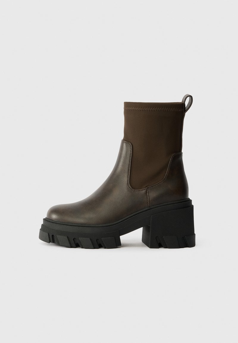 Bottine vert olive foncé avec semelle plateforme noire épaisse et talon bloc, dotée d'une languette de tirage et d'une tige en tissu extensible avec une tige lisse.