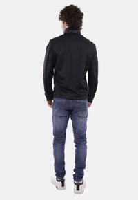 Homme aux cheveux bouclés portant une veste noire, un jean bleu et des baskets blanches, debout de dos sur un fond clair uni.