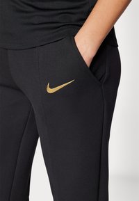 Crne atletske hlače s glatkom teksturom. Imaju zlatni Nike logo na lijevoj temi. Dizajn uključuje bočne džepove i prilagođeni oblik.