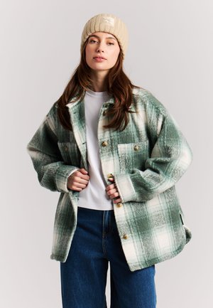 Jonge vrouw die een groen-witte geruite oversized jas draagt, wit overhemd, blauwe jeans en een beige gebreide muts tegen een effen achtergrond.