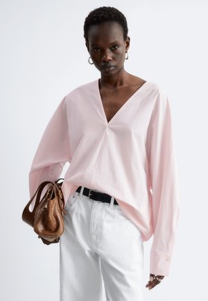 Femme portant un chemisier ample rose clair à col en V, pantalon blanc, tenant un sac à main en cuir marron, avec des cheveux courts et des boucles d’oreilles créoles argentées.