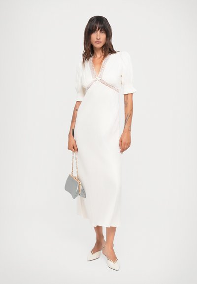 Witte midi-jurk met pofmouwen en kantdetails bij de halslijn. Gecombineerd met een lichtgrijze clutch en witte schoenen met puntige neuzen.