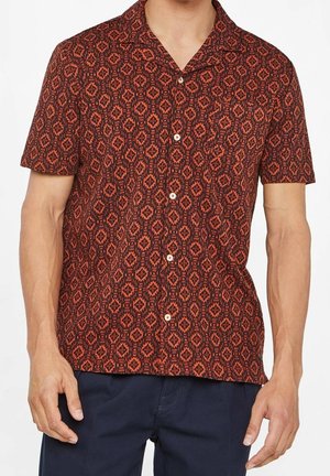 Homme portant une chemise à manches courtes à boutons avec un motif géométrique rouge et foncé, assortie à un pantalon bleu foncé, debout les bras détendus.