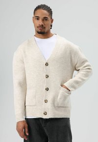 Beige gebreid vest met een V-hals, zeven bruine knopen, geribbelde manchetten en twee voorzakken. Zachte textuur en een ontspannende pasvorm.