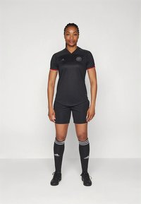 Melnā futbola kreklā ar sarkaniem akcentiem, komplektā ar melnām šortiem un ceļgalu garuma zeķēm ar baltām svītrām. Izgatavots no sintētiska materiāla.