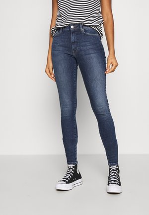Femme portant un jean skinny bleu foncé, des baskets montantes noires et un haut sans manches rayé noir et blanc devant un fond uni.