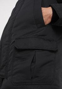 Svart isolerad jacka med en framficka med lock, synlig sömnad och en slät, matt yta.