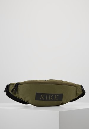 Sac banane Nike vert olive avec sangle noire réglable et double fermeture éclair, posé sur une surface blanche unie.