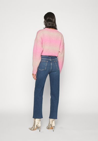 Maglione lavorato a maglia in gradiente di rosa, abbinato a jeans in denim blu a vita alta. Il modello indossa stivaletti a punta con tacco a stiletto e motivo geometrico.