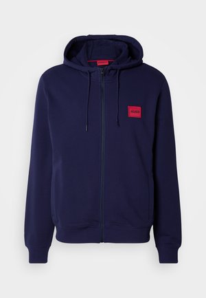 Marinblå zip-up hoodie med en mjuk textur, prydd med en röd logotyp-patch på bröstet, justerbar huva med snörning, samt ribbade avslut och ärmslut.