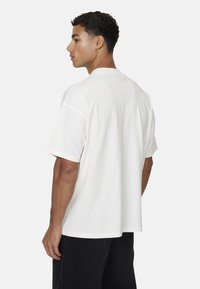 Biała bawełniana koszulka z luźnym krojem, krótkimi rękawami i okrągłym dekoltem, noszona z czarnymi spodniami. Gładka tekstura, minimalistyczny design.